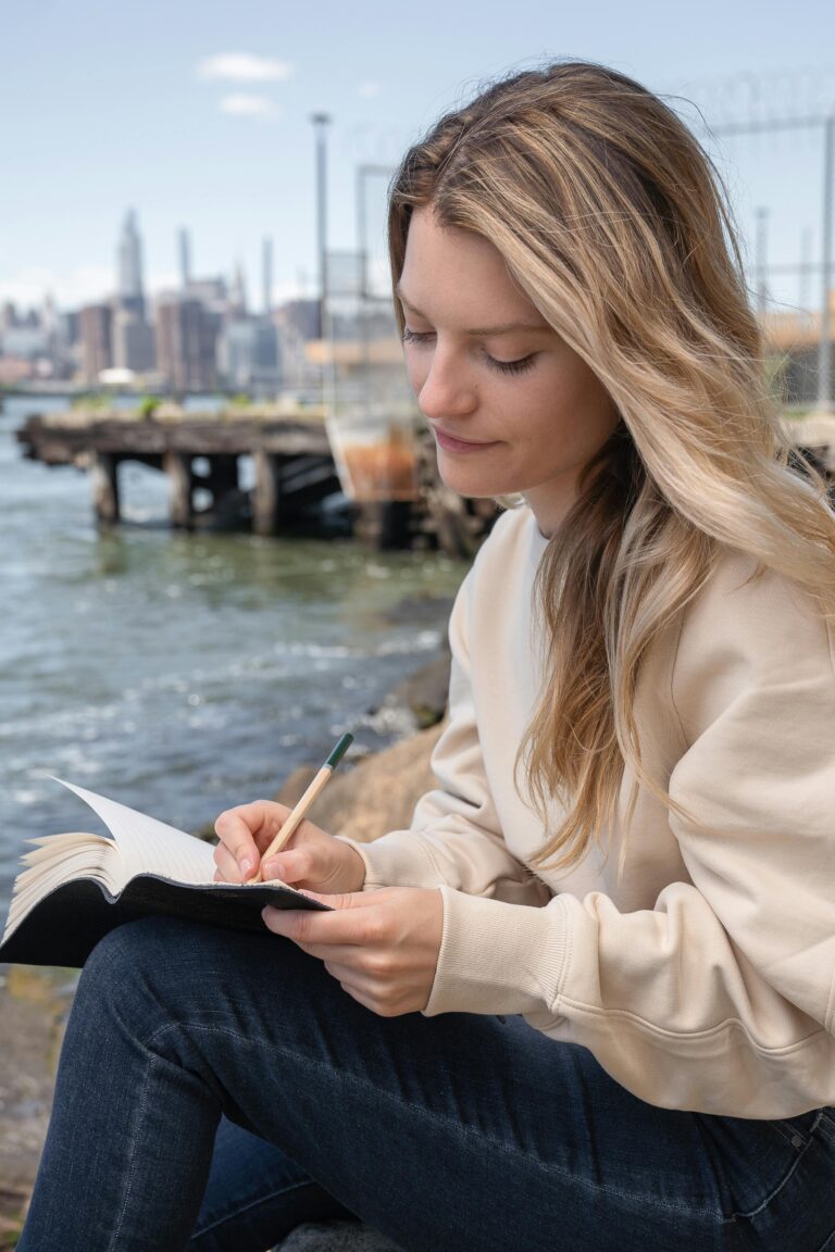 A young woman sitting by the waterfront, writing in a notebook, with a city skyline in the background.